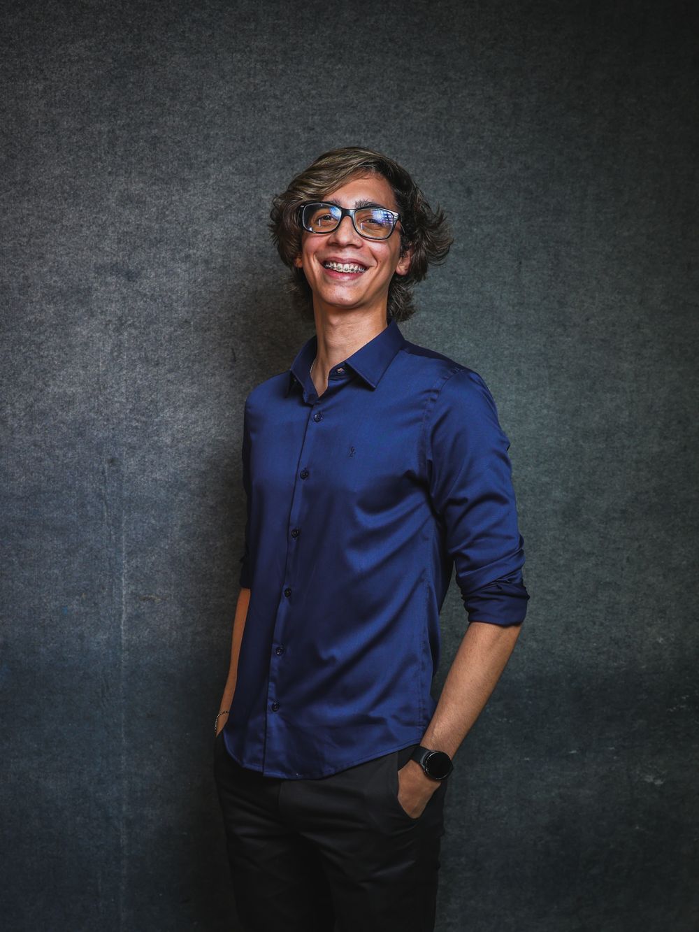 Caio Duarte sorrindo, de camisa social azul, em fundo cinza